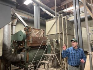 Man in baseball cap and blue plaid shirt points at industrial seed cleaning machine in warehouse.