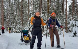 Two people nordic skiing along the All Persons Trail. One pulls a sled with a baby.