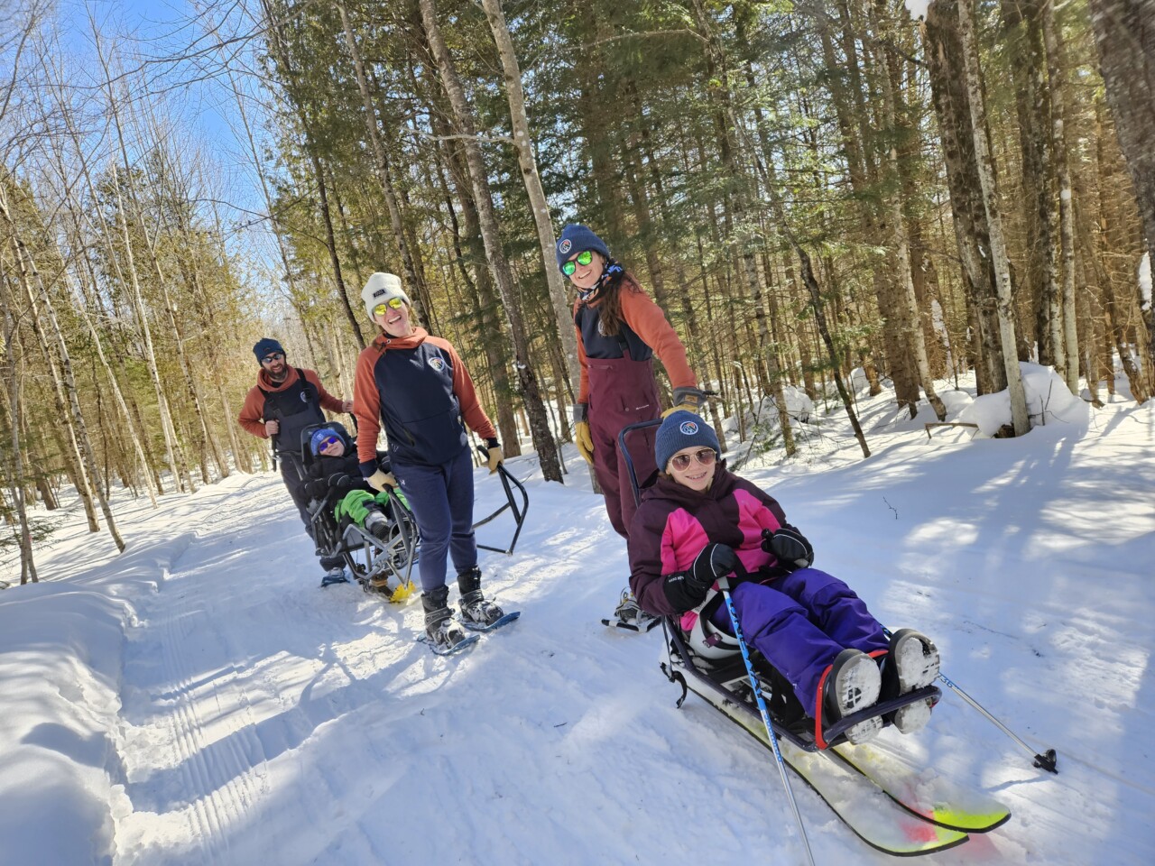 A group of smiling people stand on a snowy trail. Three are adults in snowshoes and matching jackets, two are smiling children in colorful clothing sitting in sit-ski assistive devices.