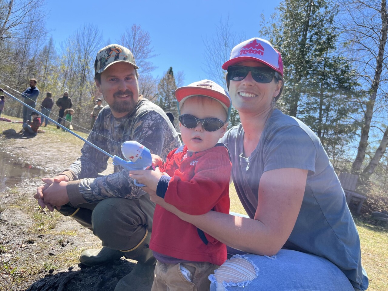 A toddler stands next to a pond holding a fishing pole with her mother and father.